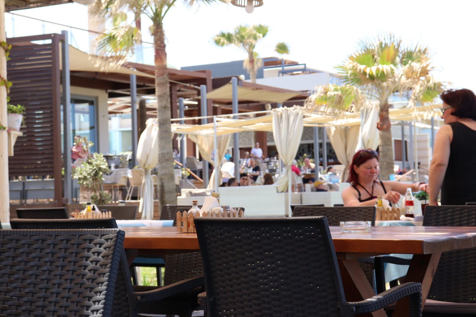 Outdoor beach café seating with palm trees and relaxed visitors in Ayia Marina