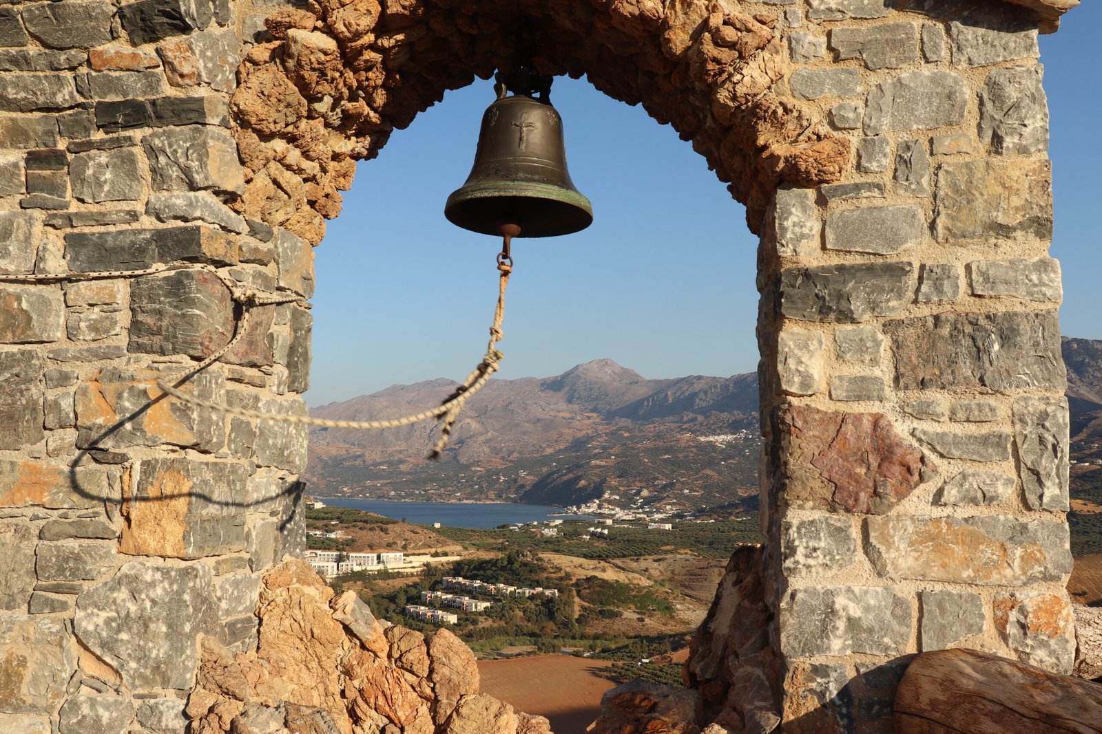 Bell of Agios Paisios Church framed by a stone arch overlooking the Plakias coastline